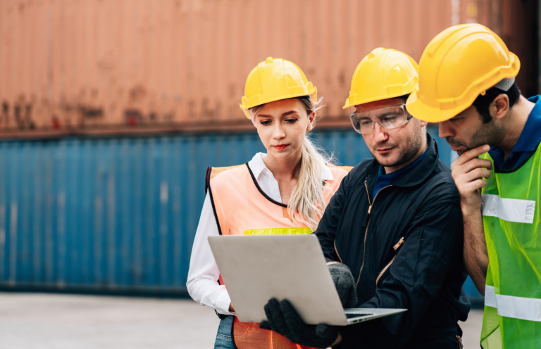 inspectores de seguridad en consultoría Tres trabajadores con cascos amarillos revisan un ordenador portátil en un entorno industrial.