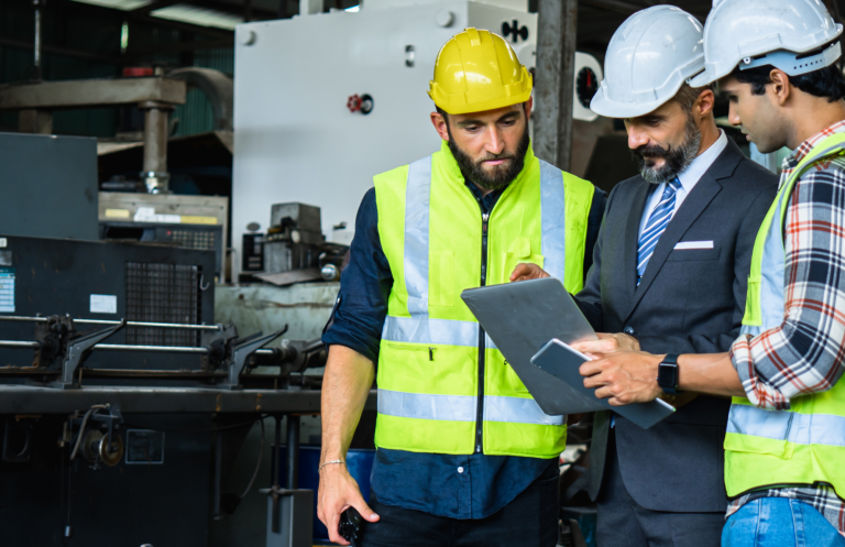 Revisión documental en consultoria Tres trabajadores en un entorno industrial revisan un documento en una tablet.